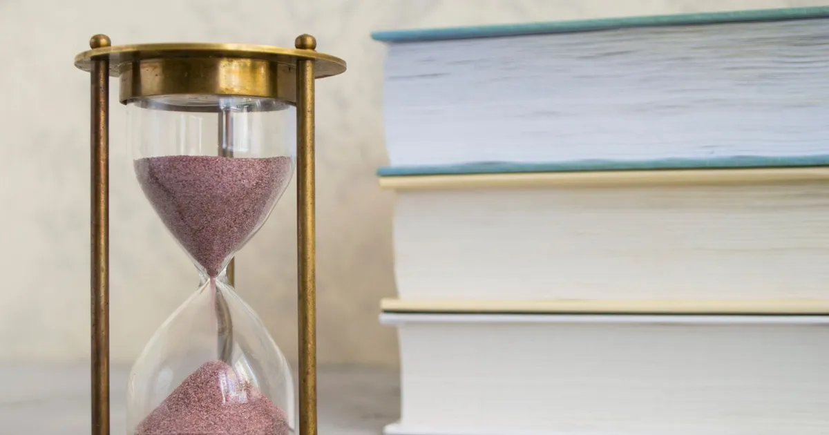 A brass hourglass with pink sand standing beside a stack of closed books