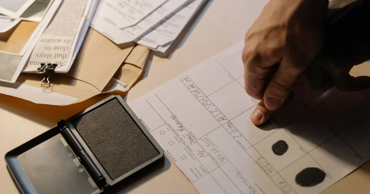 A hand being fingerprinted onto an ink fingerprint card next to case documents, representing a contractor licensing background check