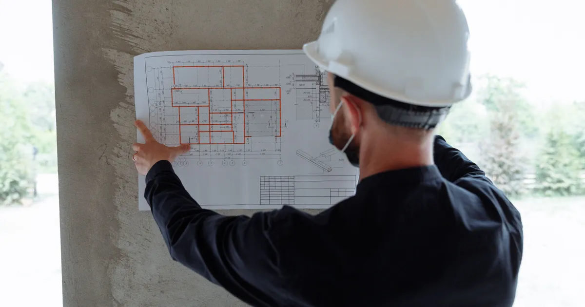 A contractor in a white hard hat studying a marked-up building blueprint on a job site