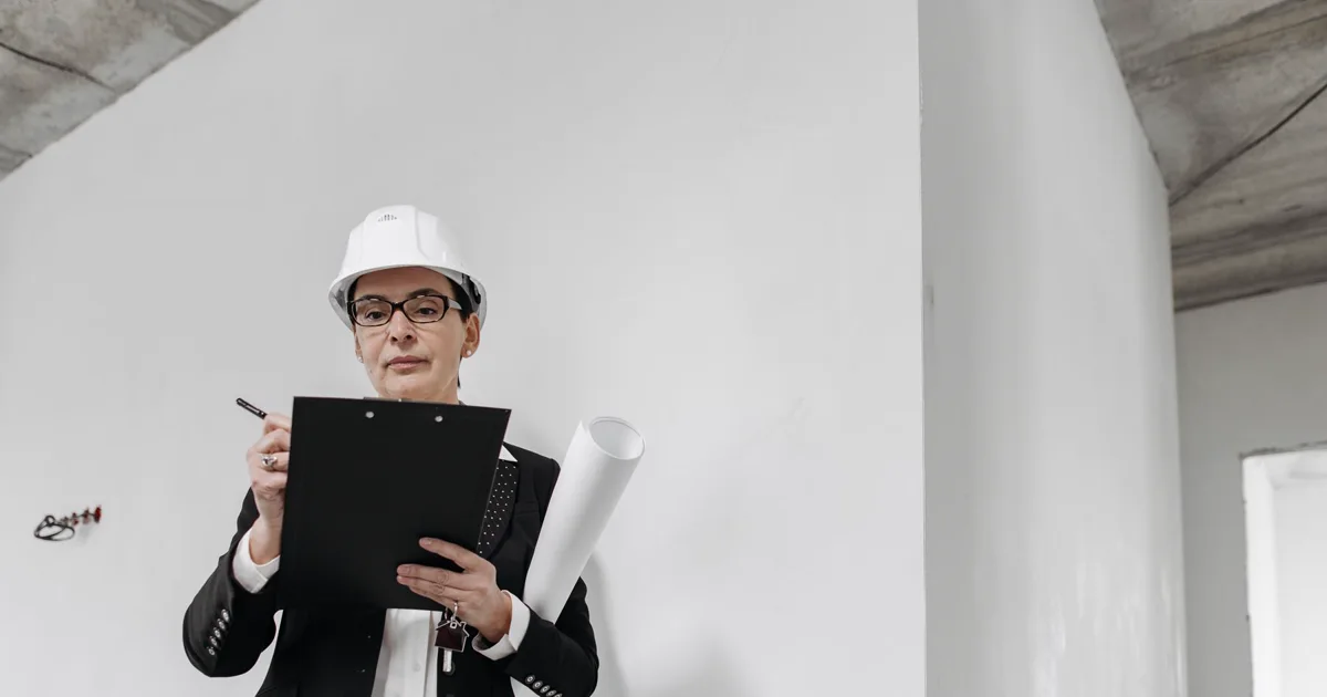 An inspector in a hard hat and business suit reviewing a clipboard and rolled blueprints inside an unfinished building